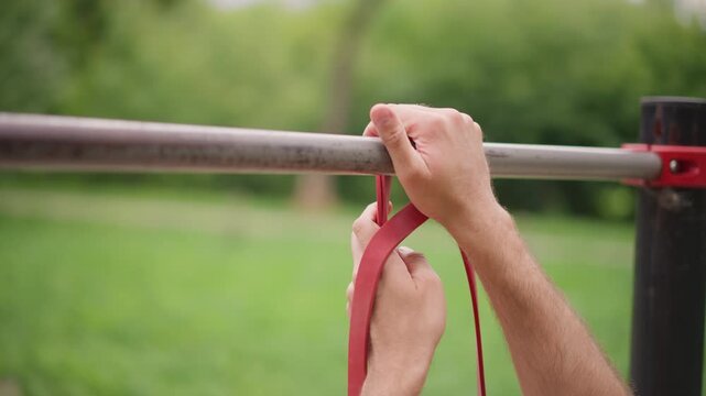 Person Securing Band On Metal Bar, Closeup Of Hands Tightening Resistance Band Onto Metal Bar At Park, Detailed Shot Of Person Securing Red Resistance Band Onto Metal Bar In Park Setting