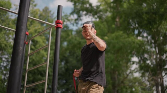 Man Adjusting Resistance Band Outdoors, Individual Attaching Resistance Band To Outdoor Bar In Park Setting, Person Preparing Exercise Resistance Band On Outdoor Structure In Leafy Park Environment