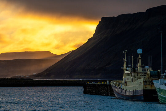View of boats moored in the serene harbor, contrasted against the imposing dark mountains under a fiery, golden sky, Grundarfjordur, West Iceland, Iceland.