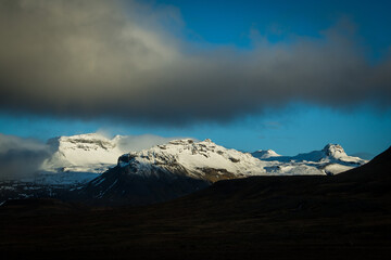 View of snow-capped mountains pierce the sky, partially veiled by dramatic clouds, casting stark shadows over the rugged landscape, Grundarfjordur, Iceland.