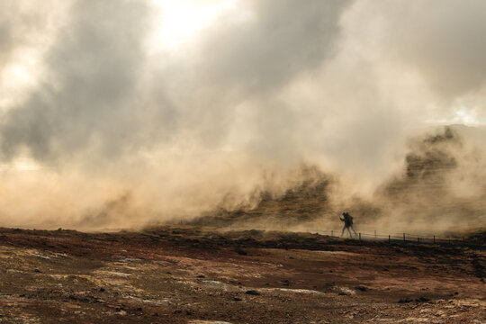 View of geothermal steam rises from the reddish-brown earth, enveloping a distant figure in a misty haze, creating an ethereal landscape, Keflavik, Iceland.