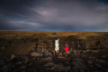 View of a solitary figure in a vibrant red cloak stands before the cascading Haifoss waterfall beneath a dramatic, brooding sky, Southern Iceland, Iceland.