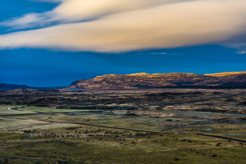 View of rugged terrain stretching towards distant cliffs under a dramatic sky, with a long road cutting through the landscape, Southern Iceland, Iceland.