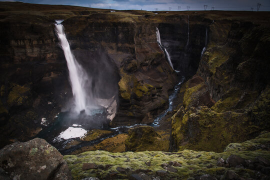 View of two powerful waterfalls plunge into a dark, craggy canyon, framed by moss-covered rocks and autumnal grasses under a moody sky, Southern Iceland, Iceland.