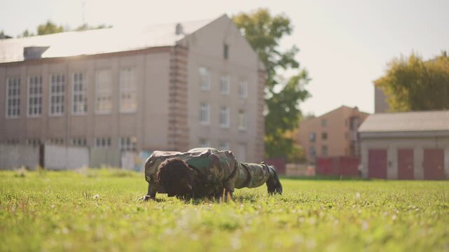 Soldier Doing Pushups On Grass In Urban Courtyard, Wearing Camouflage Uniform And Boots, Focused Fitness Drill At Morning Sunrise With Brick School Buildings In Background, Disciplined Strength
