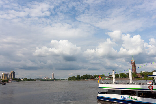 A wide view of the Rhine river in Cologne, Germany, featuring a white excursion boat in the foreground and a city skyline under a dramatic cloudy sky