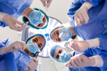 Low angle view of a diverse team of surgeons and nurses in blue scrubs, masks, and caps looking...