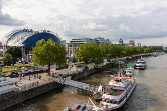 A high-angle view of the Rhine river embankment in Cologne, Germany, showing tourist boats at the pier, the Musical Dome theater, and the Great St. Martin Church in the distance under a cloudy sky