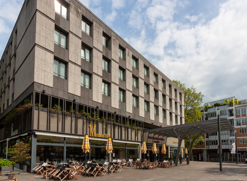 Exterior view of a modern hotel building with an outdoor restaurant terrace and yellow umbrellas in Cologne city center, Germany