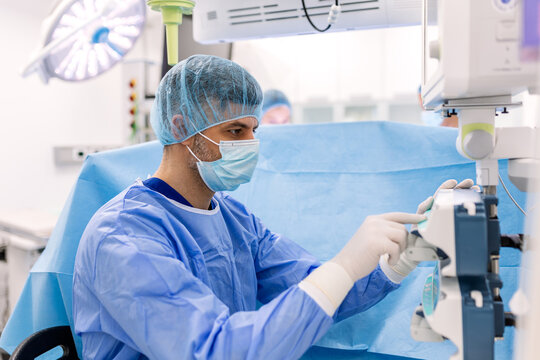Professional anesthesiologist in blue scrubs and mask monitoring patient vital signs on a screen during a surgical procedure in a high-tech hospital operating room.
