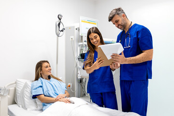 Obraz premium Smiling medical team of doctors in blue scrubs discussing patient records on a clipboard next to a young female patient lying in a hospital bed in a modern clinical room with medical equipment.