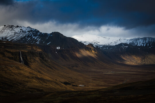 View of rugged, snow-capped mountains meet a vast, sun-kissed valley under a dramatic sky, creating a stark contrast of light and shadow, Grundarfjordur, Iceland. - Powered by Adobe