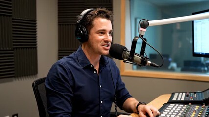 Male broadcaster speaking into microphone, wearing headphones, operating sound mixer in sound booth