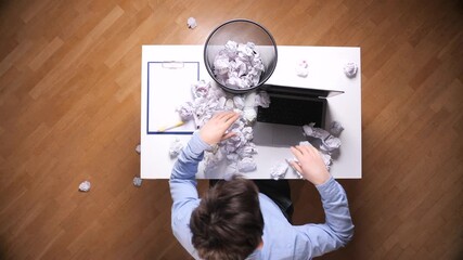 Man at cluttered office desk crumpling paper in frustration, battling writer's block and exhaustion while laptop, documents and waste bin show failed ideas and stress