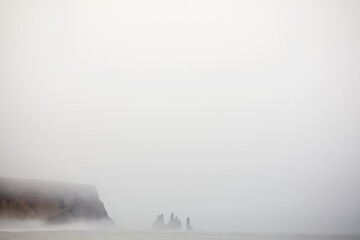View of mist-shrouded cliffs and jagged sea stacks emerge eerily from the turbulent ocean, disappearing into the thick fog, Reynisfjara Beach, Myrdalshreppur, Iceland.