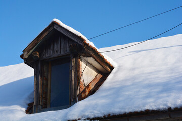 An old wooden dormer window with weathered textures protruding from a thick layer of white snow on a house roof under a clear blue sky