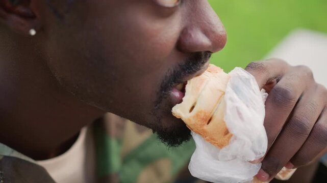 Black Soldier Eats, Individual In Uniform With Sandwich In Park, Close View Of Soldier Savoring Lunch On Park Bench, Soldier Takes Slow Bite Of Sandwich Seated On Bench Surrounded By Greenery