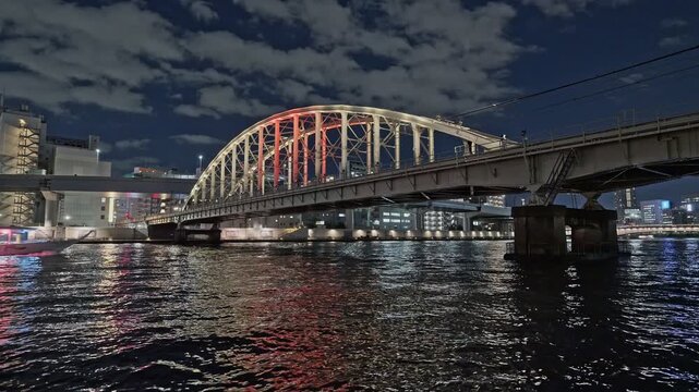 A vibrant night scene of a glowing pink sightseeing boat passing along the Sumida River, with the illuminated Sobu Line bridge in the background.