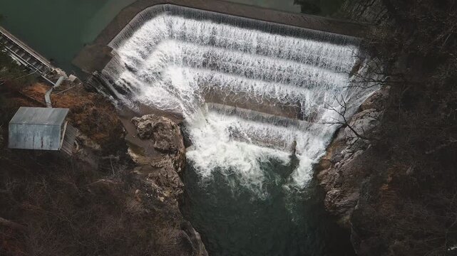 Drone glides down to Lechfall dam's tiered spillway in Fussen, capturing powerful water flow into gorge pool surrounded by autumn foliage and rocks. Dynamic hydro reveal.