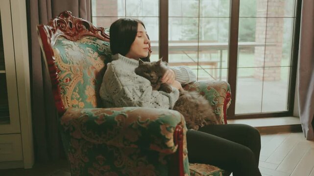 Happy woman hugging cute fluffy gray funny cat emotions of love care. Adult beauty girl smiling face at home in cozy room casual knitted clothes kisses muzzle pet holds in hands sits chair by window