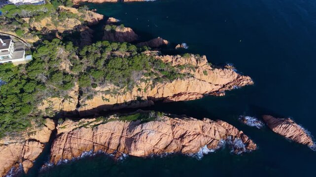 Aerial drone shot slowly flying over a beautiful rocky coastline with red cliffs and green pine trees, showing the deep blue mediterranean sea on a sunny day in sant feliu de guixols, spain