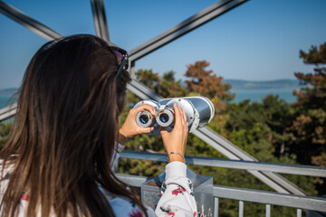 Woman looking through binoculars at Xantos viewpoint in Balatonboglar with panoramic views of Lake Balaton, forested hills, metal observation structure, and clear blue sky in Hungary
