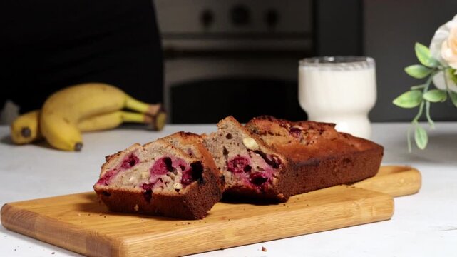 Banana bread with cherries. Freshly baked sliced banana bread with berries on a wooden board in a kitchen. Milk and bananas are on the table, adding to the cooking environment for a morning scene