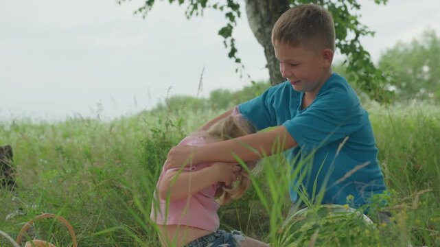 white brother helping little sister field, gentle assist in tall grass and wildflowers caring older sibling kneels to lift younger girl, hands clasped, warm expression of trust and compassion, rural
