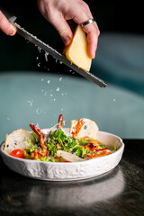 Close-up of a chef's hand grating fresh Parmesan cheese over a vibrant, healthy shrimp Caesar salad with crisp romaine lettuce, croutons, and cherry tomatoes in a white bowl.