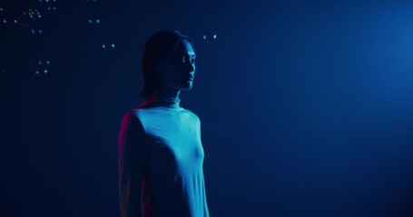 A woman stands in the blue studio light with a serious look. She starts to catch soap bubbles falling from above with her finger © m-art