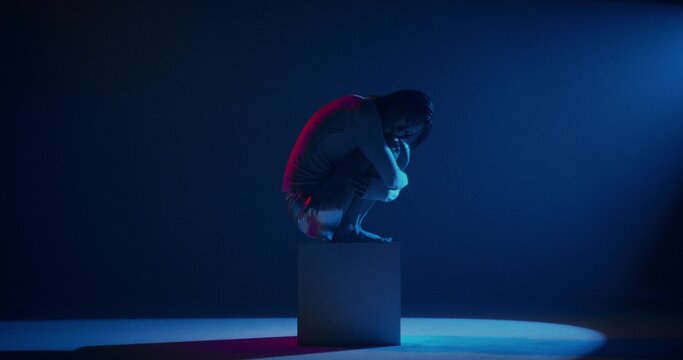 Portrait of a woman sitting on a cube in a studio. She hugs her legs. She looks at the camera with a serious look.