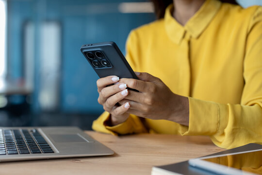 Part of the body of a young woman sitting in the office at a table with a laptop and wearing a yellow shirt, hands holding a phone