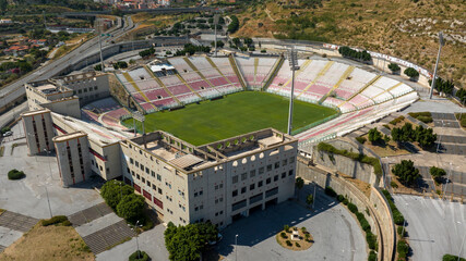 Aerial view of a large football stadium on a sunny morning. The pitch features a green pitch surrounded by white and red seating and grandstand. It is the soccer stadium of Messina, in Sicily, Italy.