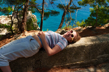 Happy young woman laughing while lying on a stone wall by the sea, joyful summer vacation moment.