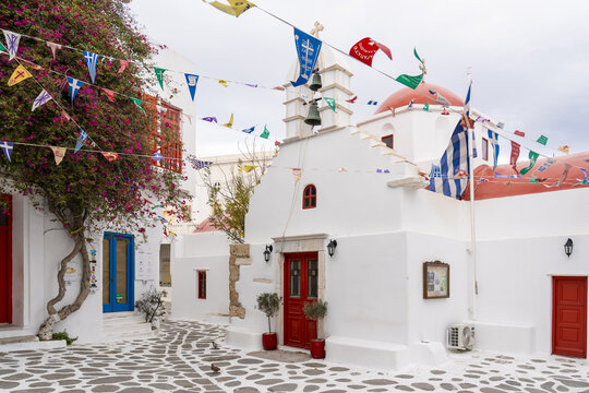 View of a traditional Greek church with whitewashed walls, a vibrant red dome, and colorful flags strung overhead on a charming cobblestone street, Mykonos, Greece.