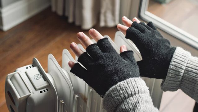 Close-up of a person's hands wearing a wool cardigan and black wooly fingerless gloves close to a domestic radiator trying to keep warm in the winter