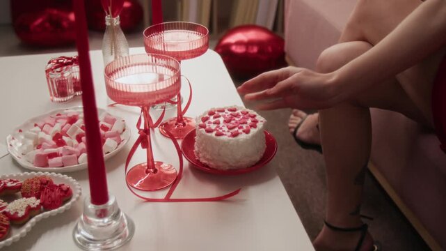 Zoom in shot of unrecognisable woman putting festive cake decorated with heart-shaped sprinkles on white coffee table set with sweets, marshmallows, bowl glasses and candles
