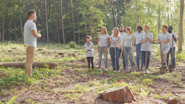 Volunteer group standing in forest area. Team of people holding shovels and tools. Caucasian male leader holding tablet device. Explaining planting plan to team. Environmental cooperation outside.