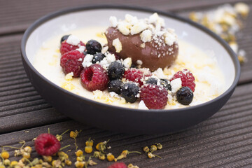 Close up of gourmet chocolate mousse dessert served with fresh raspberries, blueberries, and crushed meringue in a creamy vanilla sauce, elegantly plated on a dark rustic wooden surface.