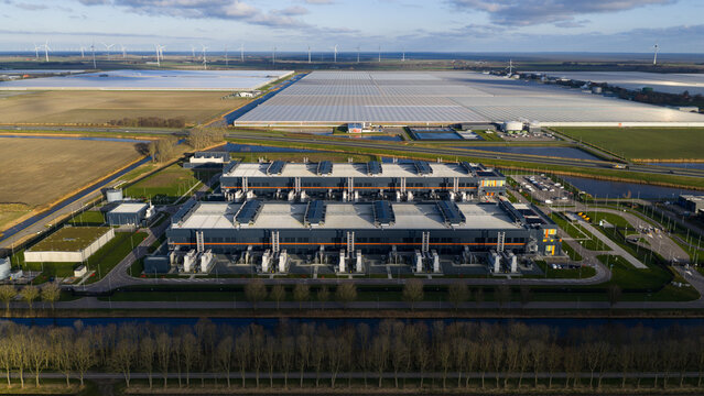 Aerial view of a vast data center contrasting with the shimmering greenhouses and distant wind turbines under the serene sky, Middenmeer, Netherlands.