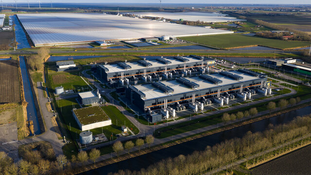 Aerial view of a vast data center's metallic structures gleaming against the soft, green fields and waterways, Data Center Agriport, Middenmeer, Netherlands.