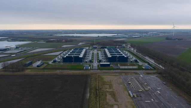 Aerial view of a vast data center complex under a cloudy sky, with wind turbines in the distance, Agriport, Middenmeer, Netherlands.