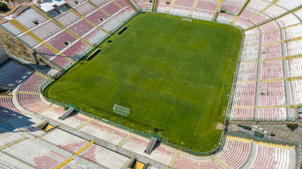 Aerial closeup of a large football stadium on a sunny morning. The pitch features a green pitch surrounded by white and red seating and grandstand.  It is the soccer stadium of Messina, Sicily, Italy.