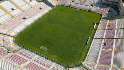 Aerial closeup of a large football stadium on a sunny morning. The pitch features a green pitch surrounded by white and red seating and grandstand.  It is the soccer stadium of Messina, Sicily, Italy.