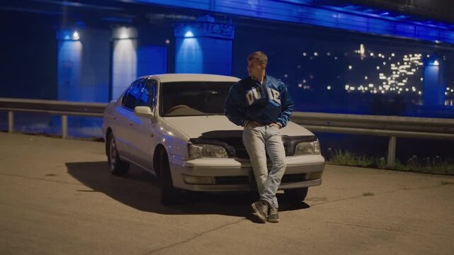 night bridge parking lot driver posing against sedan in streetwear, arms crossed, confident stance, blue underpass glow, distant traffic bokeh, editorial urban vibe, cinematic low key lighting, edgy