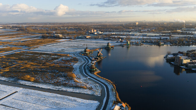 Aerial view of windmills standing proudly against the cold waters, framed by snow-dusted fields under a soft winter sky, Zaanse Schans, Netherlands.