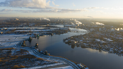 Aerial view of a serene river winding through a snow-dusted landscape, with industrial buildings puffing smoke into the sky, Zaandam, Netherlands.