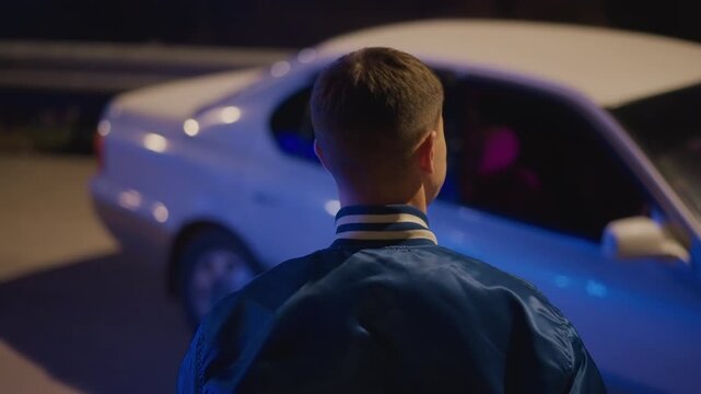 young driver walking toward car in empty lot, blue varsity jacket catching streetlight, inspects door and peers into neonlit interior, hesitates before entering cinematic framing builds anticipation