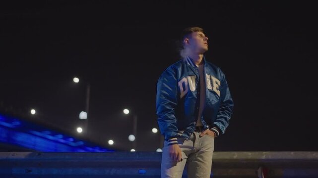 white man exhaling smoke beside bridge at night, confident stance in varsity jacket, head tilted as vapor curls upward against lit skyline, cinematic contrast and soft bokeh frames evoke street racer