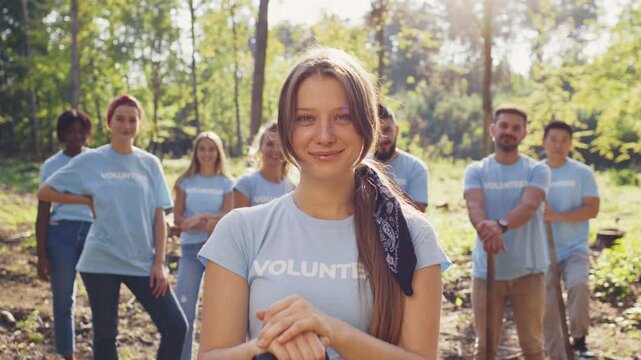 Medium view of young Caucasian female volunteer smiling calmly. Wearing team shirt and bandana in hair. Hands clasped in front. Diverse volunteers standing behind. Gathering for forest cleanup work.